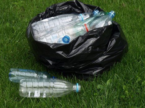 A black plastic rubbish bag sits on a grassy area, filled with multiple clear plastic bottles, some with blue or red caps, visible at the top. Several more bottles, including two with blue caps and one with a red ring, are lying on the ground nearby. The bottles appear to be PET plastic, lightweight with a smooth, glossy surface. The grass surrounding the bag is green and well-maintained, providing a natural backdrop. The scene suggests outdoor waste collection or removal, possibly on a driveway or park area, aligning with services offered by Business Waste Removal Queens Park for rubbish disposal. The lighting is natural, indicating daytime, with even illumination that highlights the transparency and textures of the plastic bottles and the crumpled appearance of the black plastic bag. Overall, the image demonstrates waste items ready for collection, emphasizing the need for professional rubbish removal in the local Queens Park area to responsibly manage waste disposal.