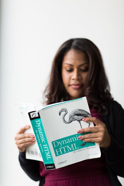 A woman with dark, shoulder-length hair and medium skin tone is holding a paperback book titled 'Dynamic HTML' by Oreilly, with a black and white illustration of a flamingo on the cover. She is dresse